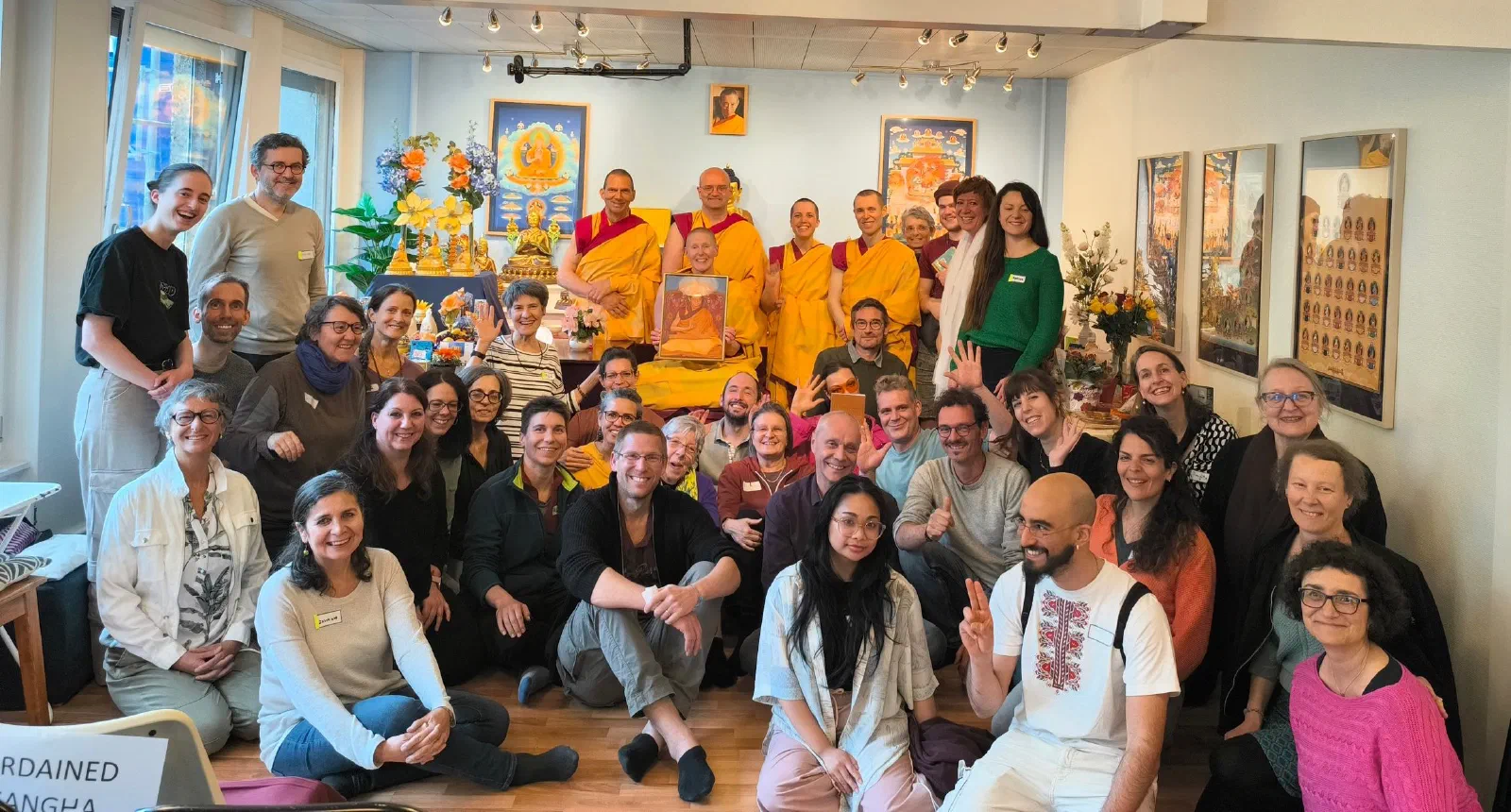 Groupe de personnes dans un cadre spirituel, incluant des moines en robe jaune et rouge, avec des décorations et des œuvres religieuses en arrière-plan, posant pour une photo collective.